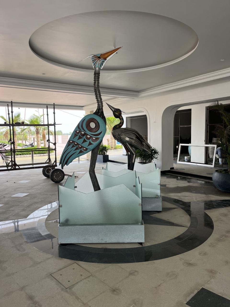 Two tall stylised heron sculptures in metal and painted panels on glass bases in a mall lobby