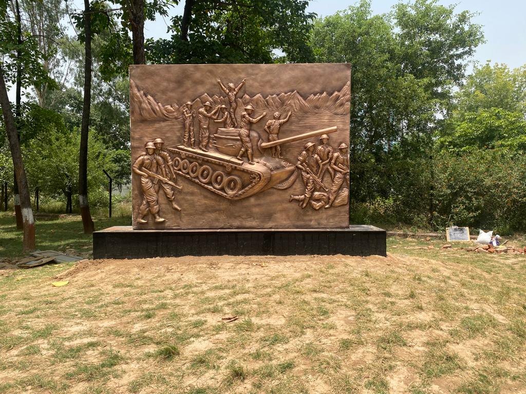 Brass-finish relief mural of soldiers celebrating on a battle tank, mounted on a plinth in a green campus
